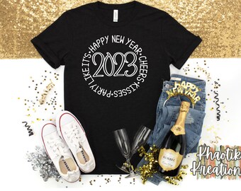 May include: A black t-shirt with a white circle design that says "Party Like It's Happy New Year. Kisses. Cheers. 2023". The shirt is laying on a white surface with gold confetti, a bottle of champagne, a pair of white sneakers, and a pair of jeans with a gold "Happy New Year" headband.