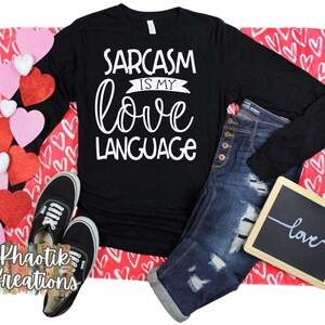 May include: Black long-sleeve shirt with white text that reads "Sarcasm is my love language". The shirt is laid out on a red background with white hearts. There is a pair of black sneakers, blue jeans, and a small chalkboard with the word "love" written on it.