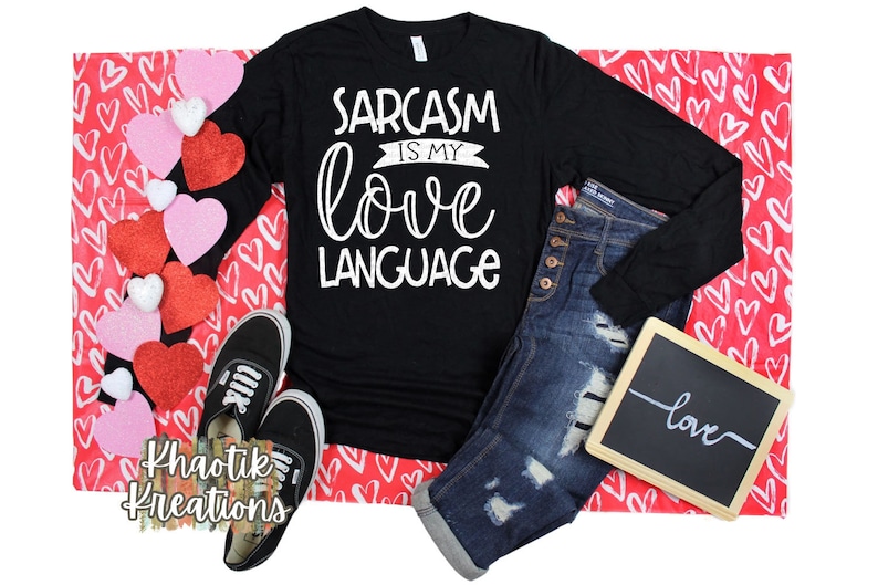 May include: Black long-sleeve shirt with white text that reads "Sarcasm is my love language". The shirt is laid out on a red background with white hearts. There is a pair of black sneakers, blue jeans, and a small chalkboard with the word "love" written on it.