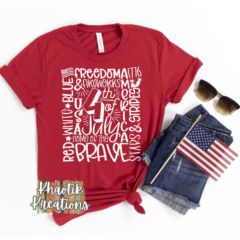 May include: A red t-shirt with a white graphic design featuring the text "Freedom," "4th of July," and "Home of the Brave." The design includes stars, stripes, and fireworks. Sunglasses, denim shorts, and a small American flag are also visible.