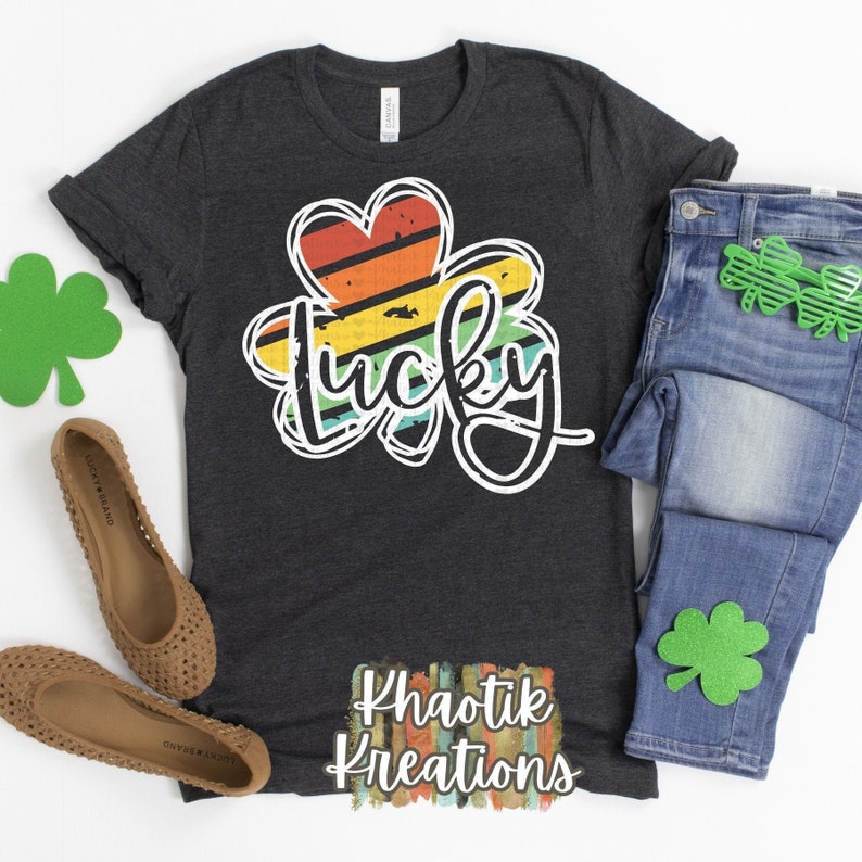 May include: A dark gray t-shirt with a white outline of a four-leaf clover with a rainbow stripe pattern inside. The word "lucky" is written in white cursive inside the clover. The text "Khaotik Kreations" is written in white with a rainbow stripe pattern below the clover.