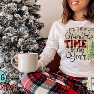 May include: A white long-sleeved shirt with the text "It's the most Wonderful Time of the Year" in black script with a red and black plaid pattern behind the word "Time". The shirt is decorated with a green Christmas tree with red and white baubles and lights.