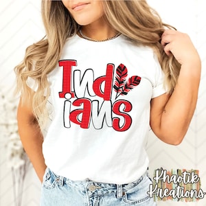 Op de afbeelding: Wit T-shirt met het woord "Indians" in rode en witte blokletters, met twee rode en zwarte veren. Het shirt heeft een ronde hals.