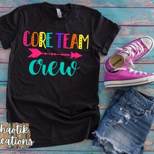 May include: Black t-shirt with the text "CORE TEAM crew" in rainbow colors with a pink arrow in the middle. The shirt is on a blue wooden background with a pair of pink Converse shoes and blue denim shorts.
