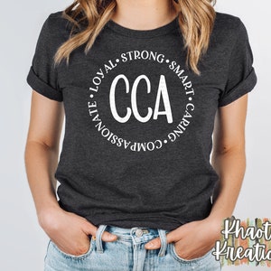 以下が含まれることがあります： 白い円のデザインが施された濃いグレーのTシャツ。円の中央には「CCA」の文字があり、周囲には「思いやりがあり、思いやりがあり、強く、賢く、忠実」という言葉が書かれています。