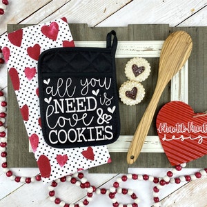 May include: Black oven mitt with white text that reads "All you need is love & cookies". The oven mitt is on a white and black kitchen towel with red hearts. There is a wooden spoon, two cookies, and a red heart-shaped sign that reads "Khaotik Kreations Design".