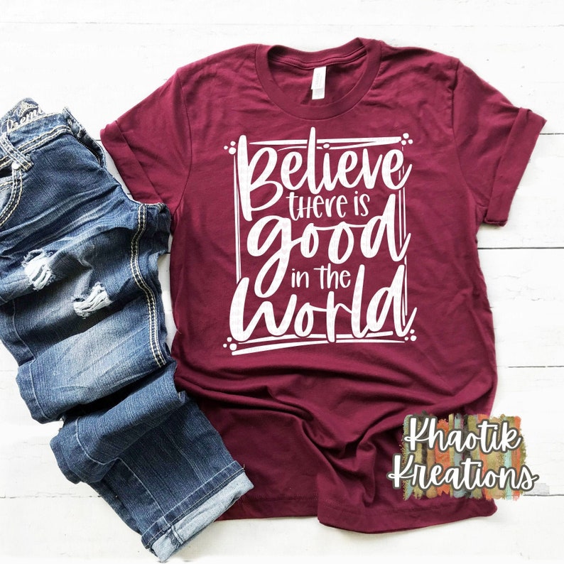 Puede incluir: Una camiseta de color granate con el texto "Believe there is good in the world" impreso en blanco. La camiseta est&aacute; doblada y colocada sobre una superficie de madera blanca.