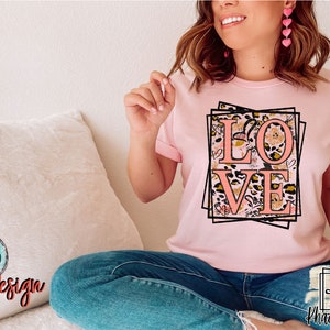Pu&ograve; includere: Una maglietta rosa con un design grafico della parola "LOVE" in lettere rosa circondata da una cornice nera e motivi floreali e animalier.