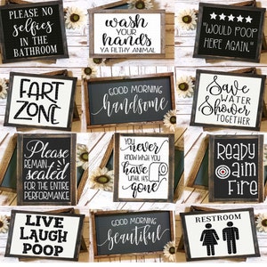 May include: A collection of framed bathroom signs with humorous phrases like "Please No Selfies" and "Would Poop Here Again." The signs feature black and white text on various backgrounds, with wooden frames and decorative sunflowers.