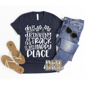 May include: A navy blue t-shirt with white text that reads "day drinking at the track is my happy place". The text is surrounded by two crossed checkered flags. The shirt is laid flat on a white surface with a pair of brown sandals and blue jeans with sunglasses on top of the shirt. The text "Khaotik Kreations" is in the bottom right corner of the image.