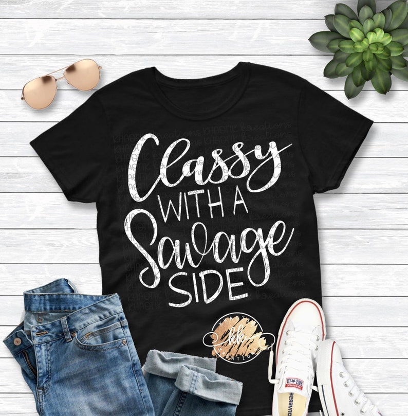 May include: Black t-shirt with the white text "Classy With A Savage Side". The shirt is paired with blue jeans, white sneakers, and sunglasses. A succulent plant is visible in the background.
