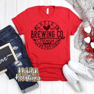 以下が含まれることがあります： ハートが描かれた円の中に「Cupid's Brewing Co. Est. 1883 Premium Love Potions」と黒文字で書かれた赤いTシャツです。