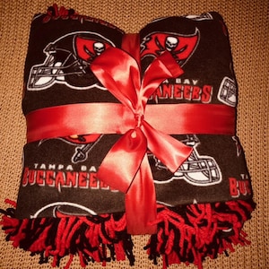 Puede incluir: Una manta de franela roja, negra y blanca con el logotipo de los Tampa Bay Buccaneers y las palabras "Tampa Bay Buccaneers". La manta está atada con una cinta roja y tiene flecos negros y rojos.