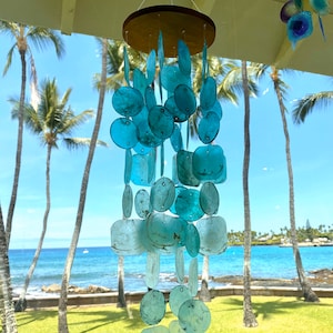 May include: A blue and white shell wind chime hanging from a wooden ceiling. The wind chime is made of many different sized shells, and it is hanging in front of a blue ocean and palm trees.