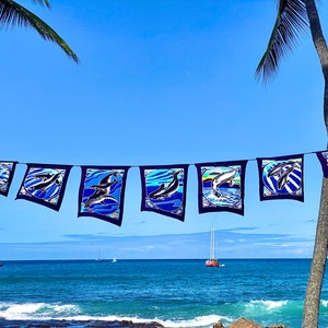 Könnte beinhalten: Eine Girlande aus blauen und schwarzen Stofffahnen mit einem weißen Walmotiv, die zwischen zwei Palmen vor einem strahlend blauen Himmel hängt. Die Fahnen hängen über einem blauen Meer mit Segelbooten in der Ferne.
