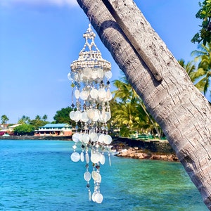 Puede incluir: Un carillón de viento decorativo hecho de conchas blancas y acentos iridiscentes cuelga de una viga de madera sujeta a una palmera. El carillón presenta elementos redondos y en forma de diamante, con un fondo de océano tropical.