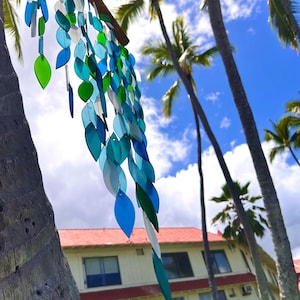 May include: A decorative wind chime featuring leaf-shaped elements in various shades of blue and green, suspended from a wooden beam. The background includes palm trees and a building with a red roof.