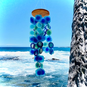 Op de afbeelding: Een handgemaakte windgong met een houten bovenkant en strengen blauwe en turquoise glazen stukken. De glazen vormen omvatten vierkanten en cirkels, wat een kustesthetiek creëert. De windgong hangt tegen een achtergrond van een blauwe lucht en de oceaan.