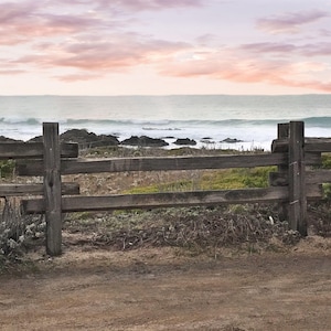 May include: A wooden fence with three horizontal rails stands in front of a view of the ocean. The ocean is a light blue color and there are white waves crashing on the shore. The sky is a light pink color with white clouds.
