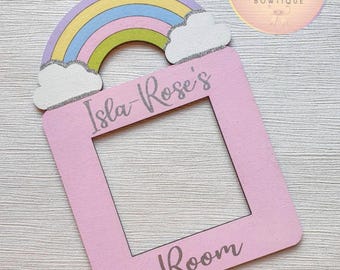 Puede incluir: Una tapa de interruptor de luz de madera rosa con un arcoíris y nubes en la parte superior. La tapa tiene el texto "Isla-Rose's Room".
