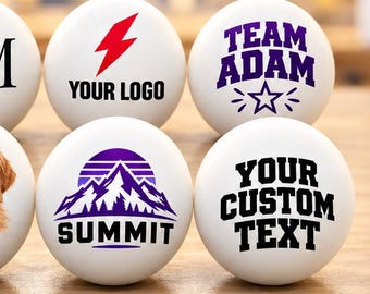 May include: Six white ping pong balls featuring custom designs including the text A&M with a heart, a red lightning bolt, the text TEAM ADAM with a star, a golden retriever portrait labeled BUDDY, a purple mountain graphic, and the text YOUR CUSTOM TEXT.
