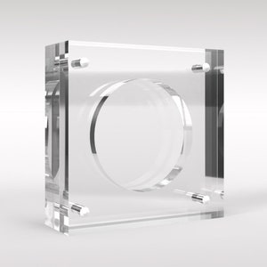 Puede incluir: Una vitrina de exhibición de monedas de acrílico transparente con una abertura circular en el centro. La vitrina tiene cuatro pequeños tornillos plateados a cada lado.