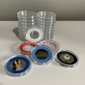 May include: A stack of clear plastic coin capsules with a white, red, blue, and black coin capsules in the foreground. The coin capsules have a clear window to display the coins. The coins feature images of Peter Rabbit, a snowman, and a penguin.
