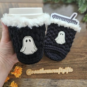 May include: Two black crochet coffee cup sleeves with white faux fur trim. Each sleeve has a white ghost applique. One sleeve is on a coffee cup. The other sleeve has a ruler sticker. The text "@southerncraftgifts" is visible.