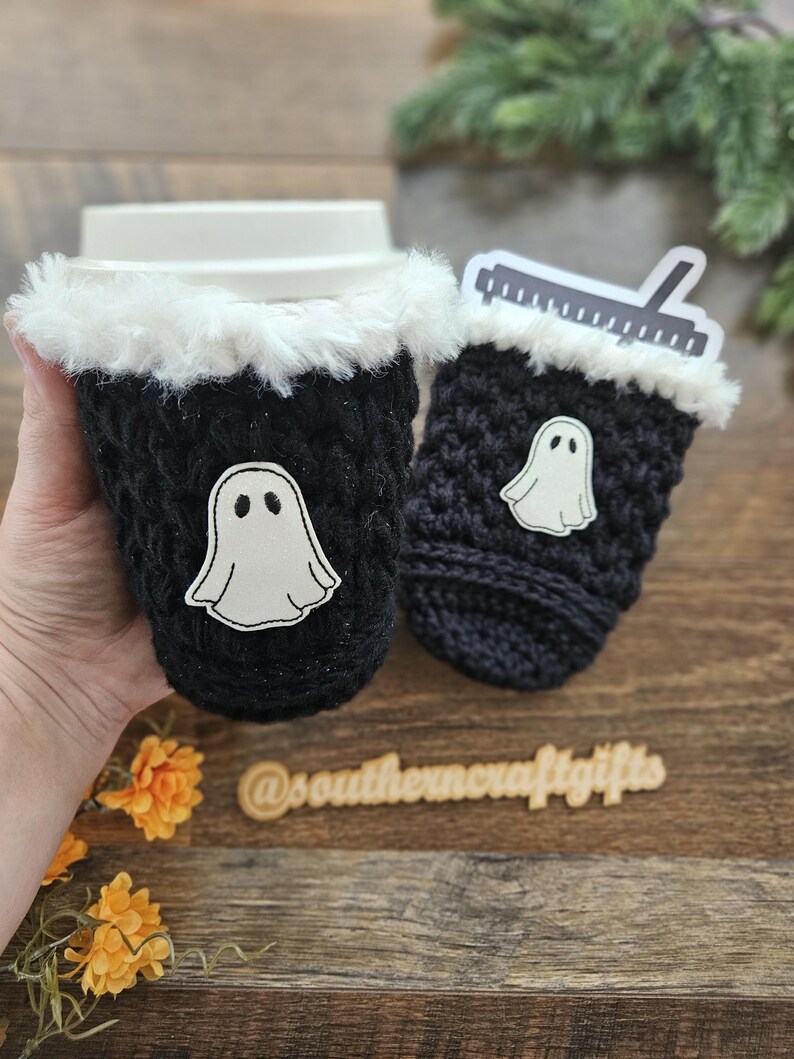 May include: Two black crochet coffee cup sleeves with white faux fur trim. Each sleeve has a white ghost applique. One sleeve is on a coffee cup. The other sleeve has a ruler sticker. The text "@southerncraftgifts" is visible.