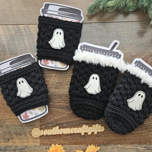 May include: Four black crochet coffee cup sleeves with white ghost appliqu&eacute;s. The sleeves are wrapped around coffee cups with fall-themed paper inserts. The cups are arranged on a wooden surface with orange flowers and greenery.