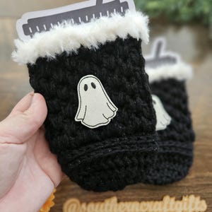 May include: Black crochet cup cozy with a white faux fur trim and a white ghost applique. A black and white paper cutout is inserted into the cozy. The cozy is designed to hold a beverage cup. The cozy is a Halloween-themed item.
