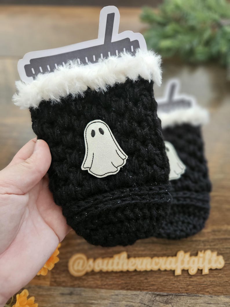 May include: Black crochet cup cozy with a white faux fur trim and a white ghost applique. A black and white paper cutout is inserted into the cozy. The cozy is designed to hold a beverage cup. The cozy is a Halloween-themed item.