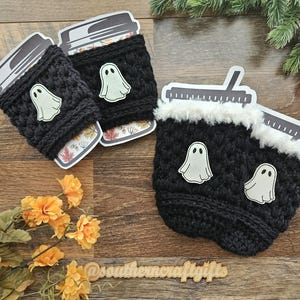 May include: Black crochet coffee cup sleeves with white ghost appliqu&eacute;s. The sleeves are decorated with a white ghost appliqu&eacute;. The coffee cups have a fall-themed design. The image also includes a wooden sign with the text "@southerncraftgifts".