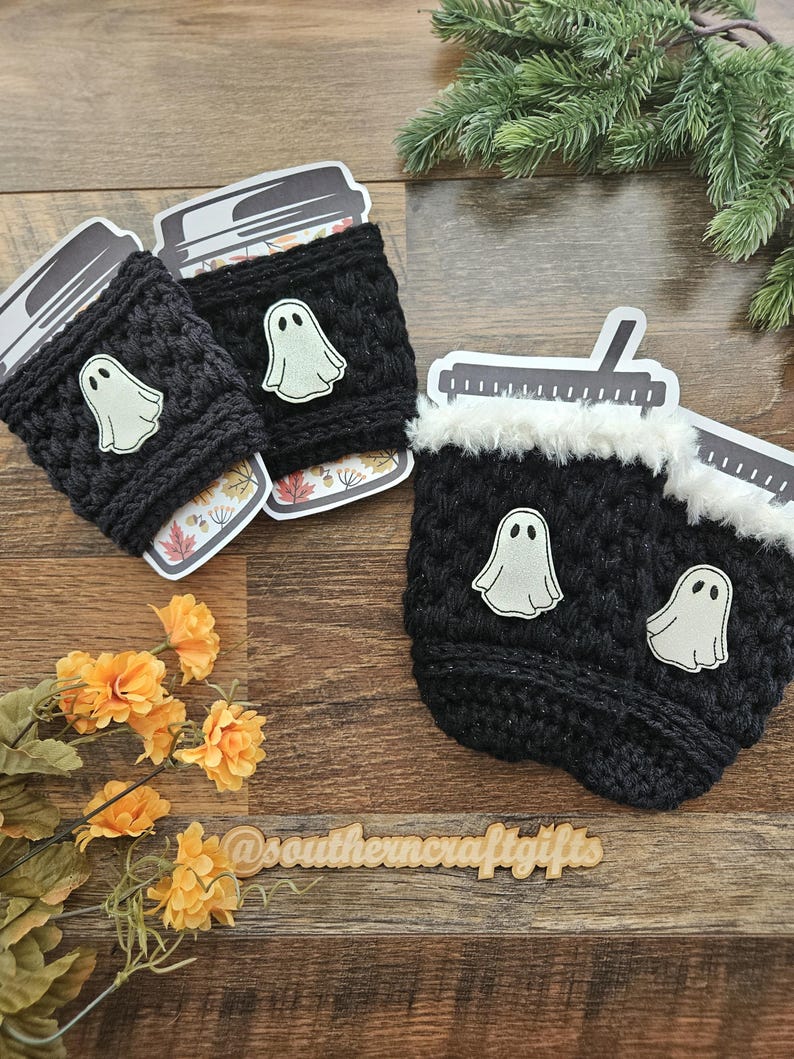 May include: Black crochet coffee cup sleeves with white ghost appliqu&eacute;s. The sleeves are decorated with a white ghost appliqu&eacute;. The coffee cups have a fall-themed design. The image also includes a wooden sign with the text "@southerncraftgifts".