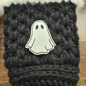 May include: A black knitted cozy with a white fluffy trim. A light gray ghost applique is centered on the front. The cozy is designed to hold a beverage container. The cozy is a Halloween-themed item.