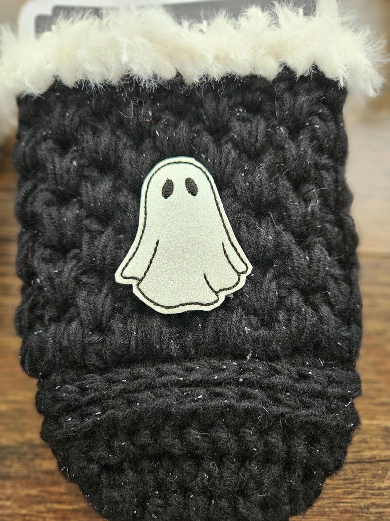 May include: A black knitted cozy with a white fluffy trim. A light gray ghost applique is centered on the front. The cozy is designed to hold a beverage container. The cozy is a Halloween-themed item.