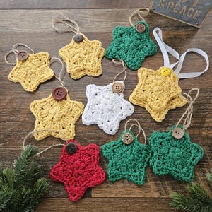 May include: A collection of handmade crochet star ornaments in various colors, including yellow, green, red, and white. Each star has a button center and a twine loop for hanging. The ornaments are displayed on a wooden surface.