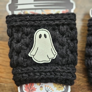 May include: A black, knitted coffee cup sleeve with a white ghost applique. The sleeve is wrapped around a disposable coffee cup with a fall-themed design. The ghost has two black eyes and a simple outline.