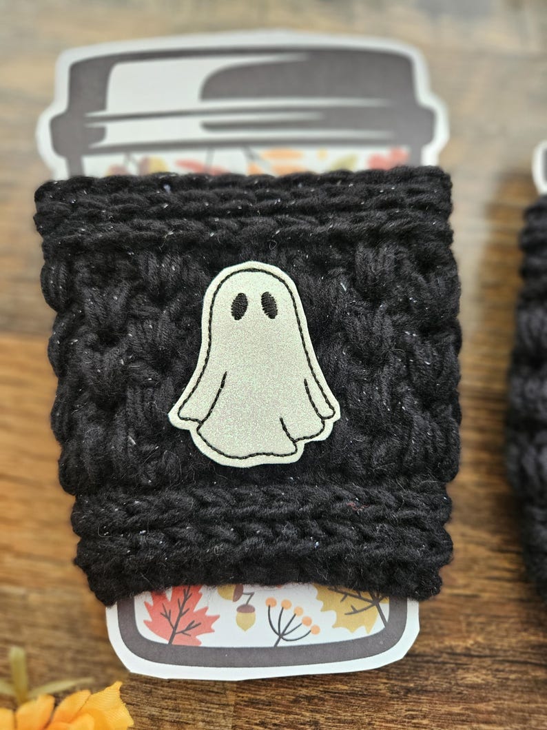 May include: A black, knitted coffee cup sleeve with a white ghost applique. The sleeve is wrapped around a disposable coffee cup with a fall-themed design. The ghost has two black eyes and a simple outline.
