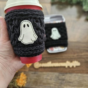 May include: A black knitted coffee cup sleeve with a white ghost applique. The sleeve is wrapped around a red coffee cup with a white lid. A second cup with a similar sleeve is in the background.