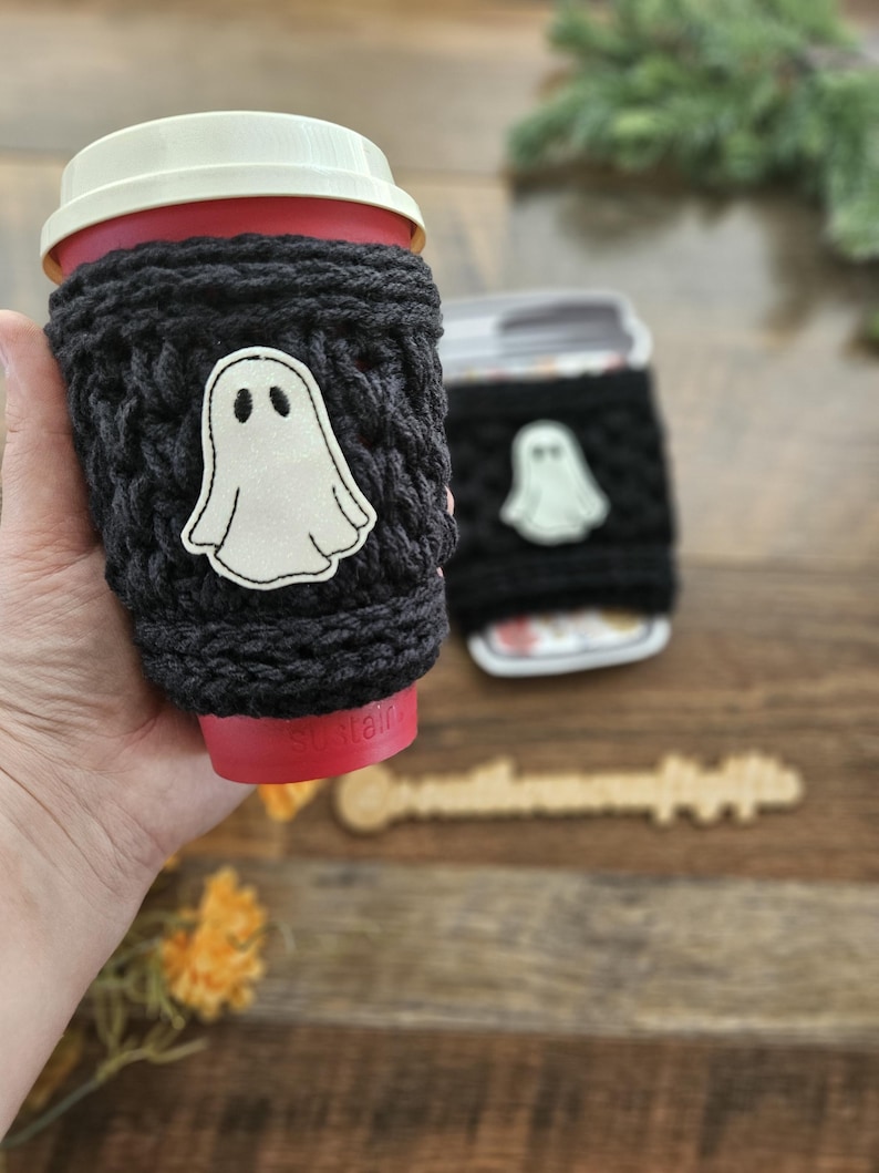 May include: A black knitted coffee cup sleeve with a white ghost applique. The sleeve is wrapped around a red coffee cup with a white lid. A second cup with a similar sleeve is in the background.