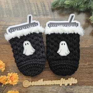 May include: Two black crocheted coffee cup sleeves with white ghost appliques. Each sleeve has a white faux fur trim and a paper lid. The sleeves are on a wooden surface with flowers and greenery.