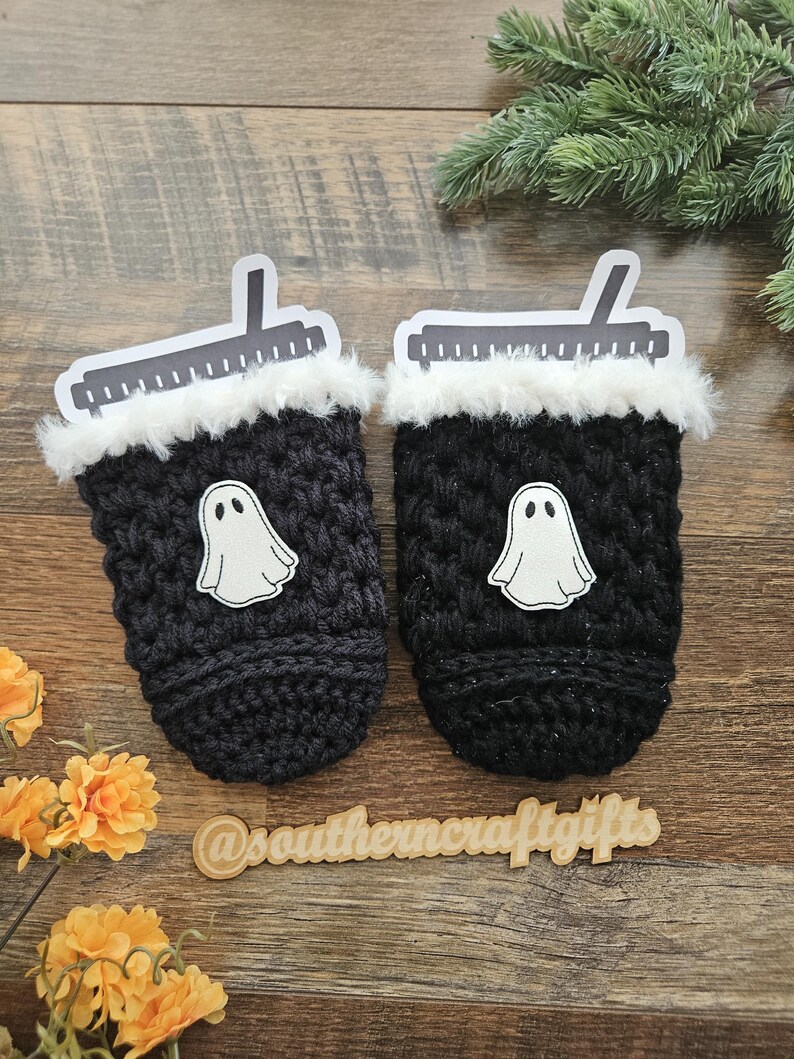 May include: Two black crocheted coffee cup sleeves with white ghost appliques. Each sleeve has a white faux fur trim and a paper lid. The sleeves are on a wooden surface with flowers and greenery.
