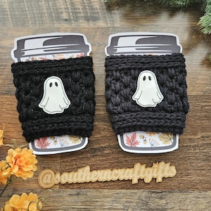 May include: Two black crochet coffee cup sleeves with white ghost appliqu&eacute;s. The sleeves are wrapped around coffee cups with fall-themed designs. The image includes the text "@southerncraftgifts".
