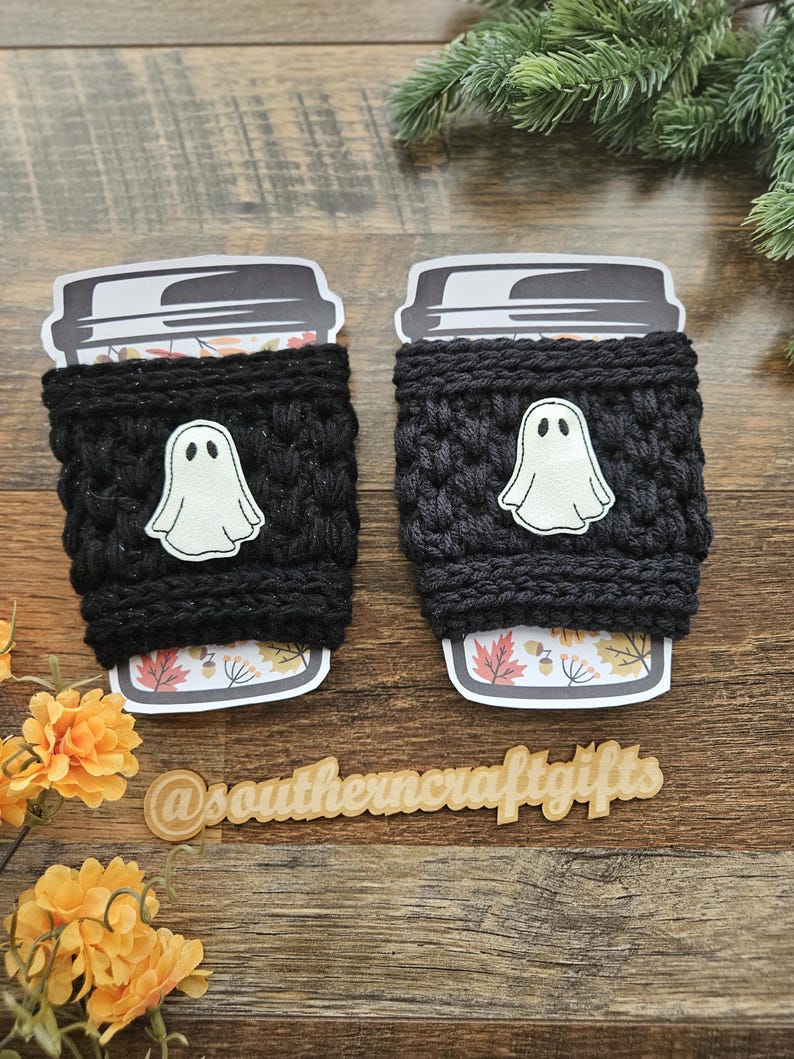 May include: Two black crochet coffee cup sleeves with white ghost appliqu&eacute;s. The sleeves are wrapped around coffee cups with fall-themed designs. The image includes the text "@southerncraftgifts".