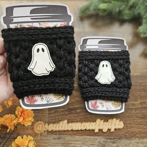 May include: Two black crochet coffee cup sleeves with a white ghost applique. The cups have a fall-themed design at the bottom. The text "@southerncraftgifts" is visible on a wooden surface.