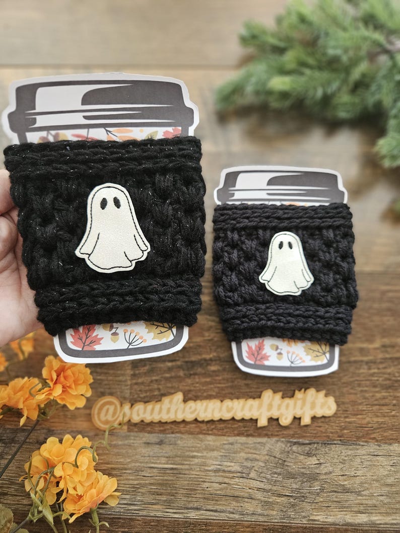 May include: Two black crochet coffee cup sleeves with a white ghost applique. The cups have a fall-themed design at the bottom. The text "@southerncraftgifts" is visible on a wooden surface.
