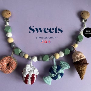 Puede incluir: Una cadena de cochecito colorida con una variedad de amuletos de ganchillo, incluyendo una dona, palomitas de maíz, caramelos y un cono de helado. La cadena está hecha con cuentas de madera y tiene clips en cada extremo. El texto "Sweets Stroller Chain" está impreso sobre un fondo morado.