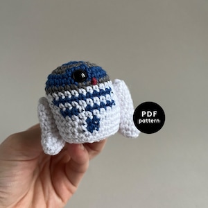 May include: A crocheted amigurumi toy resembling a character from Star Wars. The toy is white with blue and gray details and has a red nose. The toy is being held in a hand.
