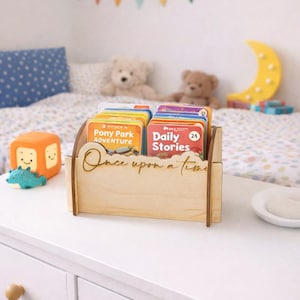 May include: A wooden book organiser with the words "Once upon a time" etched on the front. The organiser holds several children's books with colourful covers. A toy cube and a small dinosaur are on the table.
